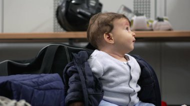 Bebek soyunma odasında oturuyor, heyecandan gülümsüyor, sıcak kıyafetlerle sarmalanmış, neşeli ve canlı bir an..