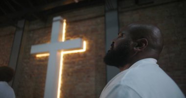 Kilise ayinine katılan kişi, modern kilise ortamında aydınlanmış haçın yanında ruhani bir yansımayla yukarı bakıyor, çeşitli cemaatlerde ibadet ediyor.