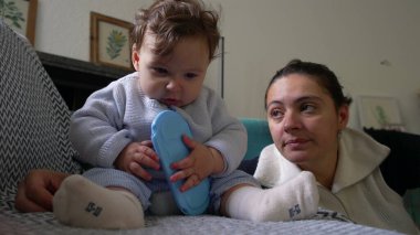 Bebek koltukta oturup mavi nesneyi çiğniyor, anne sevgiyle izliyor, kapalı alanda oyun oynuyor, bebek keşif yapıyor, aile bağı kuruyor, rahatlamış ev ortamları, çocuk gelişimi..