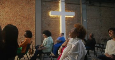 Kilise ayini sırasında şarkı söyleyen ve ibadet eden çeşitli cemaatler, arka planda aydınlatılmış haç ve tuğla duvar, maneviyat ve inancı ifade ediyor.