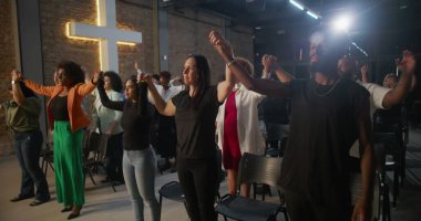 İçten bir ibadet sırasında dua ve birlik içinde el ele tutuşan cemaat, arka plandaki tuğla bir duvarda, inanç ve toplumu sembolize eden bir haç yaktı.