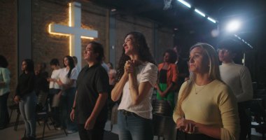 Arka planda parlayan bir haçla Hıristiyan kilisesi etkinliğine katılan çeşitli insanlar ellerini kaldırarak ibadet ediyorlar.