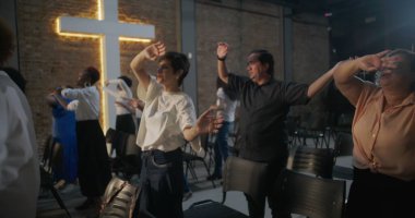 İbadet sırasında sevinçle el kaldıran ve dans eden farklı bireylerden oluşan bir grup, arka planda aydınlanan haç, canlı bir manevi bağlantı anı ve kutlama anı.
