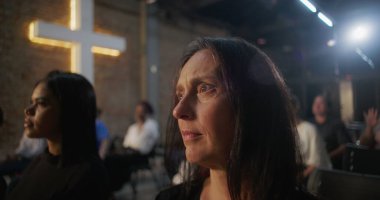 Kilisede düşünceli bir ifadeyle oturan, arka planda aydınlanmış bir haç ve cemaat olan bir kadın, kutsal cemaat toplantısındaki ibadet sırasında bunu yansıtıyor.