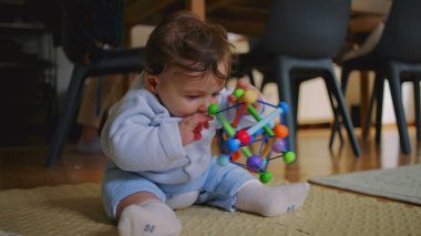Bebek yerde oturuyor, uzanıyor ve renkli bir oyuncağa dokunuyor, motor becerilerini keşfediyor ve geliştiriyor, meraklı ve ev ortamında odaklanıyor..