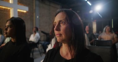 Kilisede düşünceli bir ifadeyle oturan, arka planda aydınlanmış bir haç ve cemaat olan bir kadın, kutsal cemaat toplantısındaki ibadet sırasında bunu yansıtıyor.