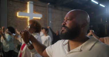 İbadet sırasında el ele tutuşan, birlik ve inanç içinde duran, arka planda toplumu ve ruhsal bağlantıyı simgeleyen haçı aydınlatan cemaat.