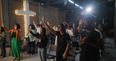 Cemaat kilise ayini sırasında el çırpıyor, arka planda haç ve tuğla duvarlar aydınlatılıyor, çeşitli topluluklar neşe ve birlik içinde.