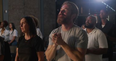 Derin dualarda çeşitli gruplar, elleri ön planda birbirine kenetlenmiş, yoğun bir bağlılık gösteren, ruhani bir toplantıda ibadet eden insanlarla çevrili insanlar.