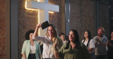 Kiliseye ibadet sırasında bağış kutusuna zarf koyan iki kadın, neşeli ve saygılı ifadeler, ruhani sunuma katılan çeşitli arka planda yer alan farklı topluluk