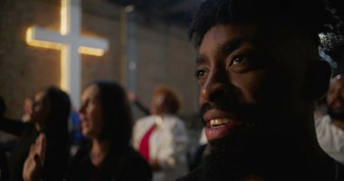 Afro-Amerikalı bir adamın kilise ayini sırasında gülümseyen, neşe ve bağlantıyı ifade eden arka planda görünen tuğla duvarda yanan bir haç profili.