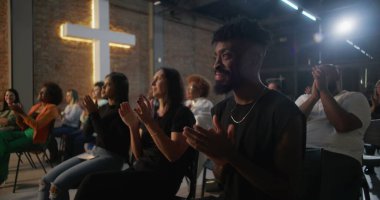 İçten bir ibadet sırasında alkış tutan cemaat, inançla ve duayla meşgul olan insanlar, tuğla bir duvarda yanan haç, ruhani bir atmosfer yaratıyor.