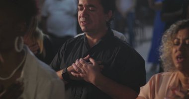 Cemaat, elleri göğsünde, gözleri kapalı, derin inanç ve ruhani bağlılıklarını ifade eden, arka planda birlikte ibadet eden farklı insan grupları, Hıristiyan ruhaniliği