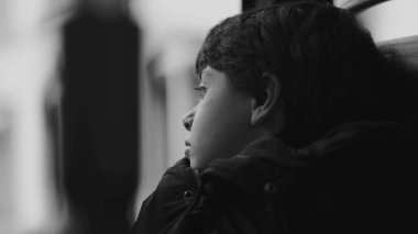 Hareket halindeki bir otobüste pencereden dışarı bakan genç bir çocuk, sessizce düşüncelere dalmış, dışarıdaki dünyayı sakin bir ifadeyle gözlemliyor, siyah ve beyaz.