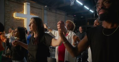 İbadet ederken el ele tutuşan, inançlarını ve bağlılıklarını ifade eden, arka planda aydınlanmış haç ruhaniliği, umudu ve yürekten birliği temsil eden bir grup insan.