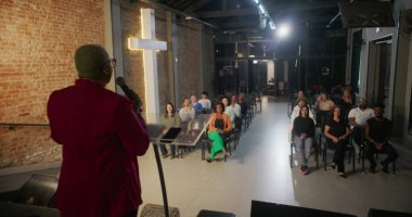 İbadet sırasında bir cemaate seslenen konuşmacı, izleyiciler sıraya dizilmiş, tuğla bir duvarda yanan haç dingin bir ruhsal atmosfer yaratıyor.