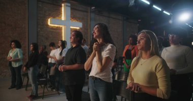 Arka planda parlayan bir haçla Hıristiyan kilisesi etkinliğine katılan çeşitli insanlar ellerini kaldırarak ibadet ediyorlar.