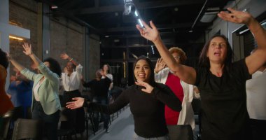 İbadet sırasında sevinçle el kaldıran ve dans eden farklı bireylerden oluşan bir grup, arka planda aydınlanan haç, canlı bir manevi bağlantı anı ve kutlama anı.