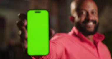 Afrika kökenli mercan gömlekli bir adamın yeşil ekran telefonuyla gülen yüz arasındaki odak noktasını şehir ortamında gece vakti açık havada gösteren bir fotoğraf.