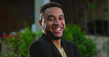 Arka planda yeşil ve dekoratif ışıklar ve şehir ortamında kollarını kavuşturarak güvenle gülümseyen Afrikalı Amerikalı bir adam.