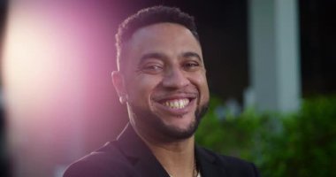 Afro-Amerikalı bir adam kameraya sıcak bir şekilde gülümsüyor, şehir ortamında yemyeşil ve yumuşak mercekli, rahat ve kendinden emin bir ifadeyle.