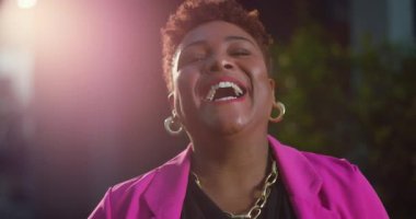 Kısa doğal saçlı, neşeli bir şekilde gülen Afro-Amerikalı bir kadın. Pembe ceketli, parlak bir gülümseme ve gün ışığında altın takılar takan bir kadın.