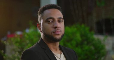 Afro-Amerikalı bir adam sakince kameraya bakıyor nötr bir ifadeyle, akşam yeşil ve bulanık dekoratif ışıklarla.