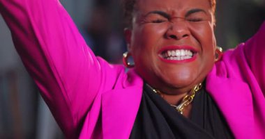 Afrikalı Amerikalı kadın kollarını kaldırıyor ve ağzı açık bağırıyor, neşe ve heyecan dolu pembe ceket ve halka küpeler içinde.