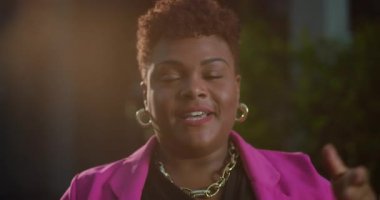 Kısa kıvırcık saçlı, kendine güvenen Afro-Amerikalı kadın kameraya doğru konuşuyor, ifade edici yüz ve canlı pembe ceket ve altın mücevher ile sıcak bir ton.