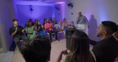 Kilisede cemaate hitap eden vaizin geniş açılı görüntüsü, plastik sandalyelerde oturan ve mesajla meşgul olan dinleyiciler.