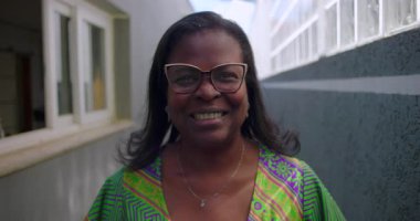 Gözlüklü ve desenli yeşil elbiseli gülümseyen Afrikalı Amerikalı kadın, gün ışığında dar bir koridorda güvenle duruyor.