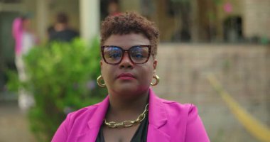 Açık havada duran pembe ceketli ve gözlüklü Afro-Amerikan kadın portresi, iddialı bir görünüm ve modern moda tarzıyla ileriye bakıyor.