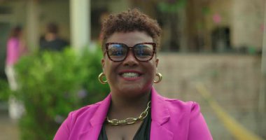 Pembe ceketli ve gözlüklü gülümseyen Afro-Amerikalı kadın kameraya neşeli bir ifadeyle bakıyor, kendine güveni ve tarzını dışa vuruyor.