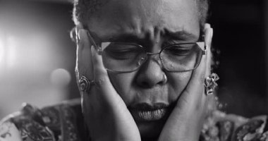 Siyah beyaz çekimde Afrika kökenli Amerikalı bir kadın derin kaşları ve kapalı gözleriyle yüzünü tutarak duygusal yoğunluğunu ve iç mücadelesini gösteriyor.