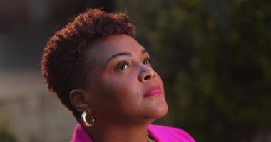 Afro-Amerikalı bir kadın umut dolu bir gülümseme ile gökyüzüne gözlerini açıyor, dışarıda sıcak ışıkta imanını ve iyimserliğini ifade ediyor, pembe ceket ve altın halkalı küpeler takıyor.