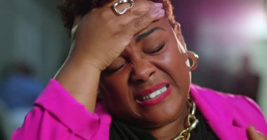 Afro-Amerikalı kadın gözlerini kapatıyor ve elleri alnında somurtuyor. Pembe ceket ve altın aksesuarlarla yorgunluk ve basıncın duygusal ifadesi.