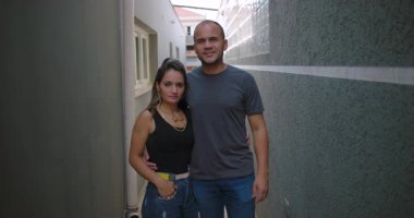 Latin erkek ve Latin kadın dar bir sokakta yan yana duruyorlar, yakın duruş ve sakin ifadeler, açık hava ortamında sıradan bir çift portresi.