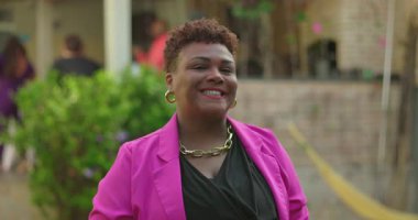 Afro-Amerikalı bir kadın, pembe ceketini düzeltirken, rahat bir şekilde dışarıda duruyor, kendine güvenen varlığıyla gülümsüyor ve sıradan bahçe düzeninde şık altın takılar takıyor.