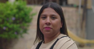 Arka bahçedeki kameraya özgüvenle bakan uzun saçlı genç Latin kadın portresi, rahat duruşu ve doğal ışığı olan sakin yüz ifadesi.