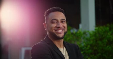 Afrika kökenli Amerikalı bir adam gece şehir ortamında gülümsüyor, kolları çapraz, rahat ve yumuşak sinema arka planında yeşillik ve merceklerin parlamasıyla ulaşılabilir.