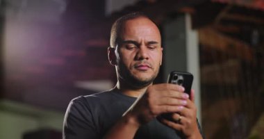 Afrika kökenli Latin kökenli bir adam akşam ışığı altında dikilirken akıllı telefon ekranına odaklanmış. Konsantrasyonunu ve ifadesiz yüz ifadelerini sergiliyor.