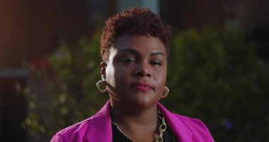 Kısa doğal saçlı Afro-Amerikalı bir kadın düşünceli bir sessizlikle dışarı bakıyor. Pembe ceket ve altın küpeler takıyor.