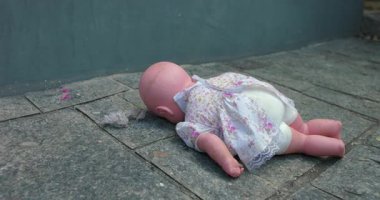 Yaşlı bebek taş kaldırımda yatıyor, eskimiş oyuncağın detaylı görüntüsü dışarıdaki duvarın yanında terk edilmiş halde.