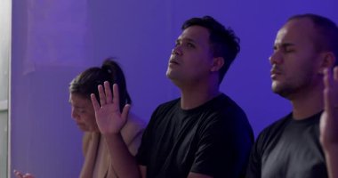 Latin erkekler ve çeşitli gruplar ellerini kaldırarak dua ediyorlar. Gözler ortak ruhani anda kapalı, sakin meditasyon ortamı.