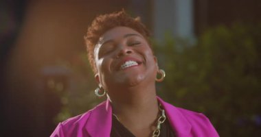 Kısa doğal saçlı, açık havada kameraya gülümseyen, parlak pembe ceketli ve halka küpeli, neşeli ve karizmatik bir yüzü olan Afro-Amerikan kadın.