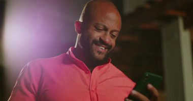 Afrika kökenli kel bir adam gece dışarıda telefon okurken gülümsüyor. Şehir ışıkları altında mercan gömlekli bir mesaja neşeli bir tepki veriyor.