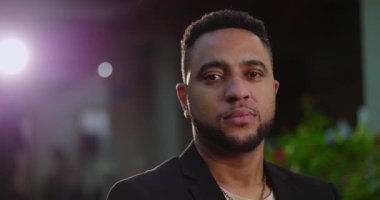 Alacakaranlıkta dışarıda dikilen Afrikalı Amerikalı adam, tarafsız bir ifadeyle doğrudan kameraya bakıyor, mimari elementlerle hafifçe aydınlatılıyor.