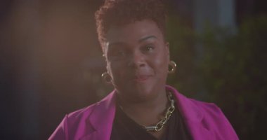 Kısa doğal saçlı Afro-Amerikalı kadın açık havada yumuşak göz teması kuruyor, pembe ceket ve altın aksesuarlar takıyor ve nazik bir ifade ve sıcaklıkla ifade ediyor.