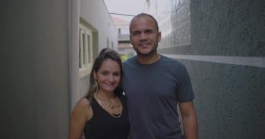 Latin erkek ve Latin kadın dar bir sokakta yan yana poz veriyorlar. Sevecen vücut diliyle gülümsüyorlar. Rahat bir çift portresi çiziyorlar.