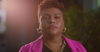 Kısa kıvırcık saçlı, açık havada kameraya ciddi ciddi bakan Afro-Amerikalı bir kadın. Pembe ceket ve altın takılar giyiyor.